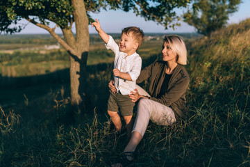happy young mother sitting on grass and looking at cute little son pointing with finger outdoors
