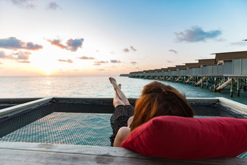Back of view woman lying on terrace looking on sunset with villas over water in Maldives feeling...