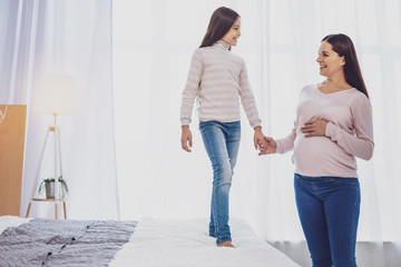 Family love. Full length of delighted long haired girl holding her pregnant adorable mother by hand while looking at her with happiness in eyes