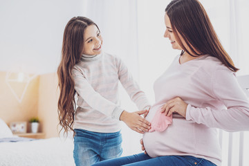 So sweet. Waist up of optimistic young pregnant woman looking at her cute daughter while showing...