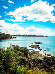 Hekerua Bay on Waiheke Island in New Zealand with sail boats on the water below and rocks and trees in the foreground. Taken on a bright Summer day.