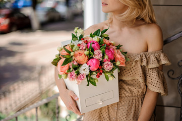 Blonde girl in beige dress with open shoulders holding a square box filled with bright flowers