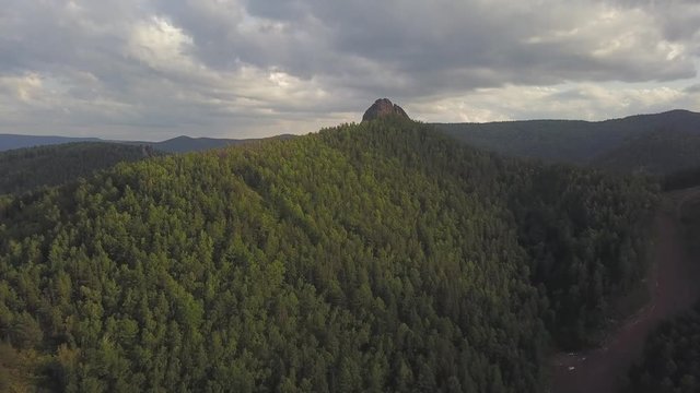  Aerial Drone Footage View: Aerial View flight over flight of high mountains covered with green coniferous pine trees and cedars. At the top of the mountain is a huge stone in the flag, set in honor o