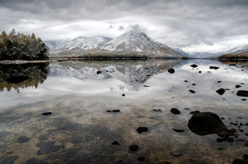 Mountain lake Froliha, pine tree and stones via transparent water with snow at mirror lake