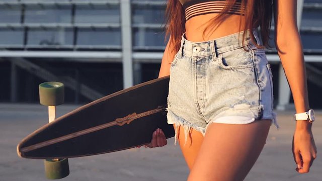 Young woman in jeans shorts holding longboar and walking. Close-up.