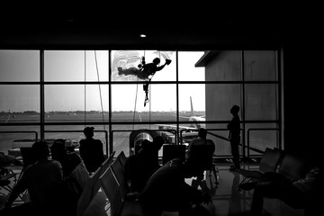 Employee is using a rope to rappelled down to clean the glass of an airport building. There are passengers waiting to board and are looking at the cleaning staff. Silhouette concept.