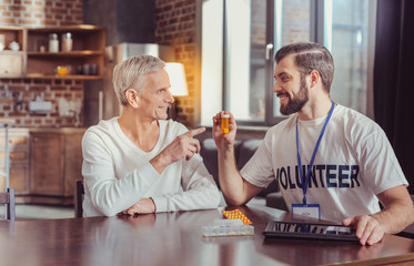Take it. Beardful pleasant sociable man sitting by the table holding pills and looking at a retired.