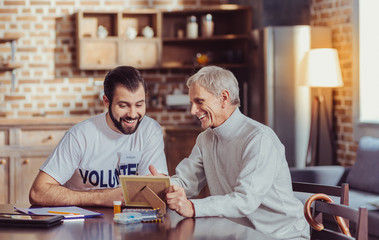 Look at this. Friendly senior satisfied man sitting by the table smiling and showing a photo to a volunteer.