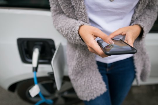 Woman Using Mobile Phone While Charging Electric Car At Charging