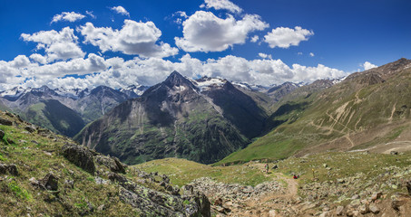 Fototapeta na wymiar Wide panoramic view overlooking the mountain peaks, glaciers and rivers flowing down, forest on the sides of mountains, hiking trails, grass meadow on sunny summer day blue sky with clouds.