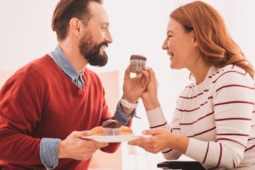 Just baked. Joyful pleasant couple eating cupcakes while expressing happiness at home