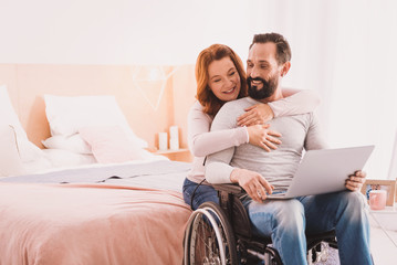Love you so much. Cheerful handsome disabled man using a laptop while his wife embracing him