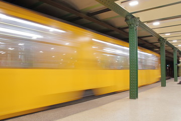 Żółty pociąg przejeżdża przez podziemną stację, skład w pędzie, rozmyty, wyraźny oświetlony peron z ozdobnymi zielonymi filarami - obrazy, fototapety, plakaty