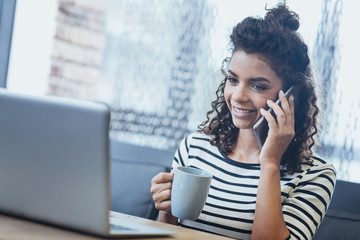 Business call. Inspired female freelancer drinking coffee while chatting on phone