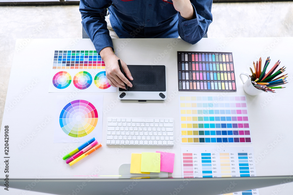 Poster Image of male creative graphic designer working on color selection and drawing on graphics tablet at workplace with work tools and accessories, top view workspace