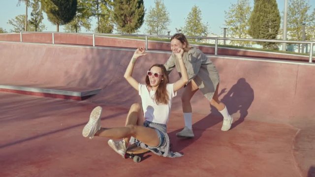 Two smiling female friends with american flag having fun with skateboard in the skate park. Woman sitting on skateboard being pushed by a friend