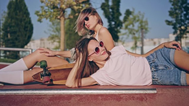 Two happy young girls with longboard having fun together while sitting at the skate park. Summer fashion, leisure and vacation concept