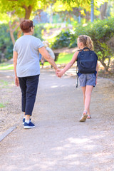 Grandma and pupil kid holding hands going to school.Little girl with school bag or satchel walking to school with grandmother. Old Parent and daughter, grandmother and granddaughter, back view.