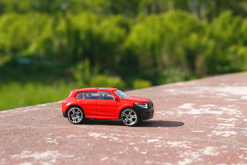 red toy car on the background of green trees, there is a car on a concrete slab in the daytime a bright sun