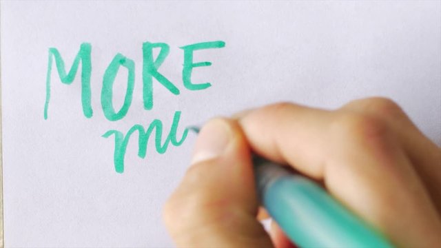 Hand writes MORE, MORE, MORE!!! on white paper with a green brush pen