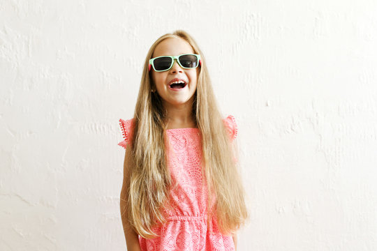 Little girl w/ long golden hair in fashionable casual outfit, wearing sunglasses & smiling, laughing, making funny faces. Five years old daughter in pink dress posing. Background, close up, copy space