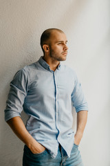 Brutal man in the blue shirt and blue jeans shorts posing holding hands in pockets in the the flat with white walls near the window in Spain