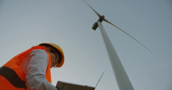 Caucasian woman in a helmet and with computer in hands checking the work of the huge windmill turbine producing energy. View from below.