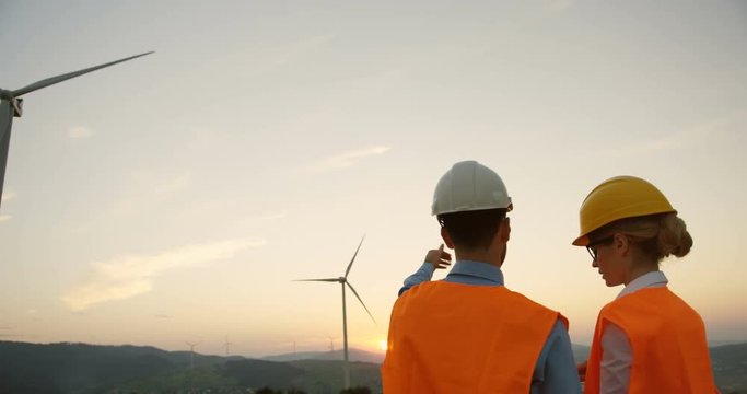 Back view of the two Caucasian male and female consultants in helmets talking about work of big windmills turbines.