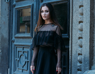 Obraz na płótnie Canvas portrait of a beautiful girl, a brunette girl in a black dress, stands near a big black door, goes into it. a new stage in life, enter
