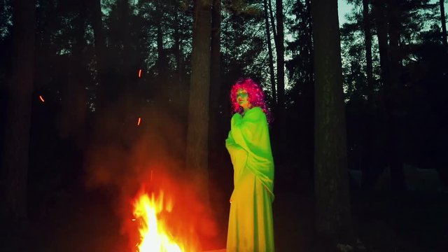 The green witch stands by the fire at night and looks around.