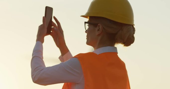 Rear of the good looking caucasian woman in the helmet and glasses taking pictures outside on the sunset at the windmills renewable energy station.