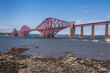 Naklejka premium Queensferry, Scotland, UK - June 14, 2012: Closeup of Red metal iconic Forth Bridge for trains over Firth of Forth between blue sky and blue water. Rocky shore up front.