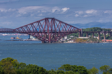 Queensferry, Scotland, UK - June 14, 2012: One of three segments of Red metal iconic Forth Bridge over Firth of Forth between blue sky and water. Buildings on other side of water. Green foliage.