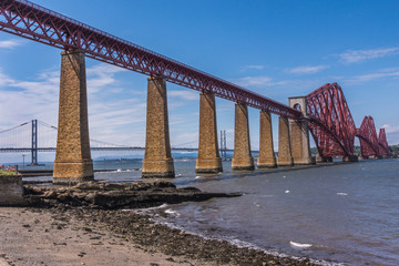 Queensferry, Scotland, UK - June 14, 2012:  Two bridges, Road suspension bridge and  Red metal iconic Forth Bridge over Firth of Forth between blue sky and water. Rocky shoreline up front.