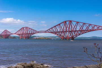 Queensferry, Scotland, UK - June 14, 2012: Closeup of Red metal iconic Forth Bridge for trains over Firth of Forth between blue sky and blue water. Old battery buildings on other side.
