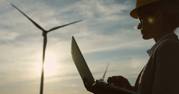 Close up of the shadow silhouette of Caucasian woman engineer in the helmet and glasses working on the laptop while standing outside with the huge windmill tourbine on the background.
