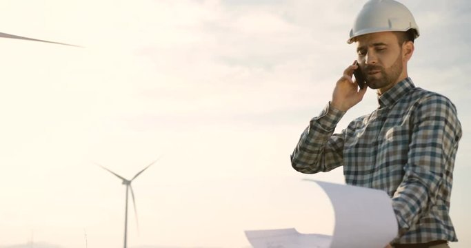 Caucasian male worker of the windmill power station in a helmet speaking on his phone and holding some documents or drawings in his hand.