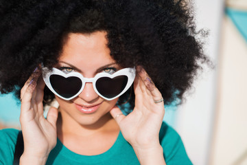 Sensual portrait of young pretty woman with african curly hairstyle and heart-shaped sunglasses. Beautiful girl with green eyes smiling to camera at summer outdoors.