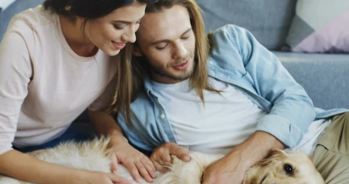 Portrait of the young Caucasian good looking couple sitting on the floor while hugging and caessing their labrador dog. Close up.
