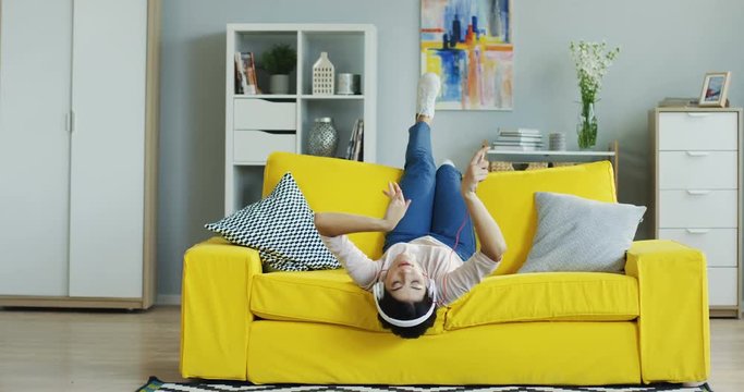 Joyful young woman lying upside down on the ellow couch in the living room and listening to the music through headphones and smartphone.