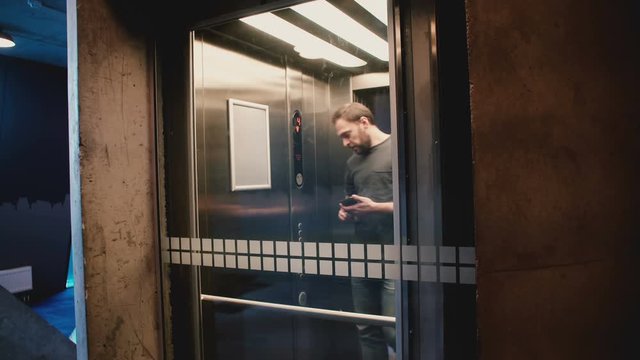 Young happy man enters elevator, pushes the button, door closes and he rides up using smartphone mobile office app.