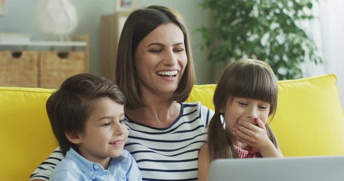 Smiled pretty Caucasian mother with two cute kids sitting on the yellow couch, laughing and watching something on the laptop.