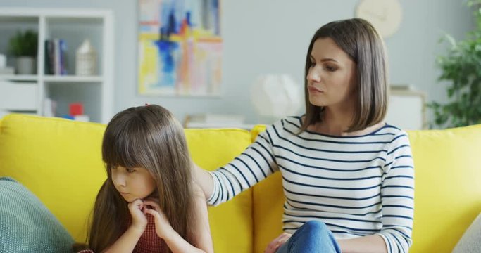 Cute Caucasian little girl sitting on the couch in the living room sadly and her pretty young mother caressing her and trying to pity.