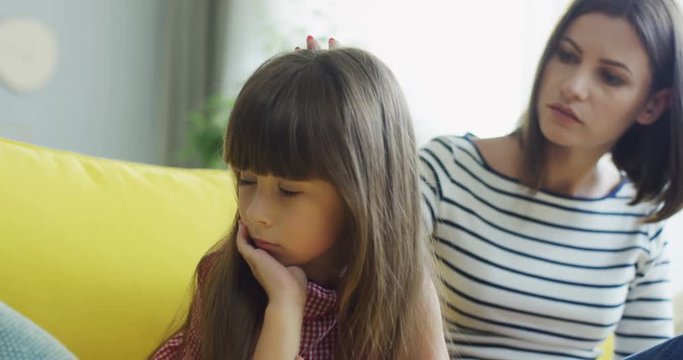 Close up of the upset small girl and her mother caressing her and talking to calm her down. At home.