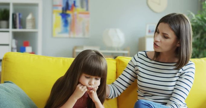 Attractive woman caressing her small cute daughter because she being upset and not in the good mood. On the yellow couch at home.
