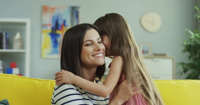 Portrait shot of the Caucasian young mother and little daughter sharing secrets and whispering on the ear in the living room.