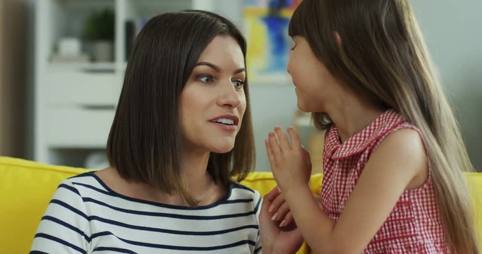 Cute small girl and her beautiful young mother gossiping and whispering while sitting on the sofa in the living room.