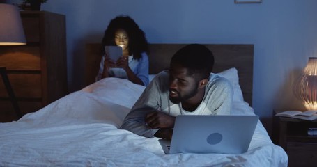 Young African American man lying on the bed and watching something on the laptop computer late in the evening. Her smiled girlfriend with tablet computer sitting on the background. - Powered by Adobe