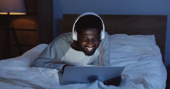 Atractive young African American man in headphones listening to the music or watching a video while lying on the unmade bed in late evening.