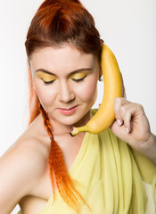 Young redhead woman holding banana near her face like smartphone on a white background.